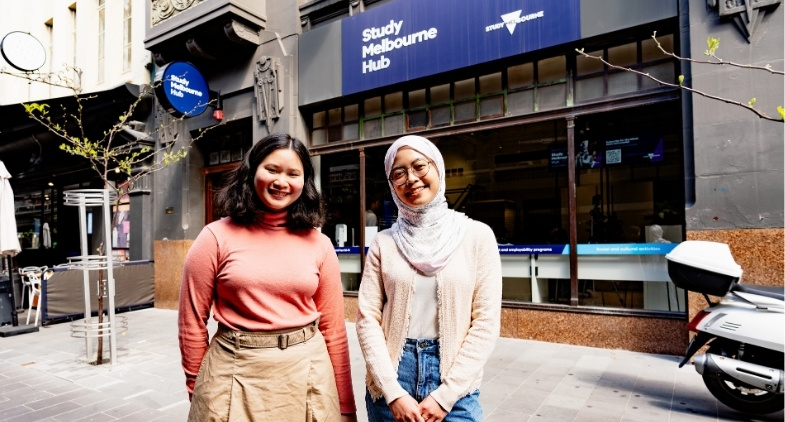 Two students smiling in front of the Study Melbourne hub with signage, heritage building, awning and carpark in background