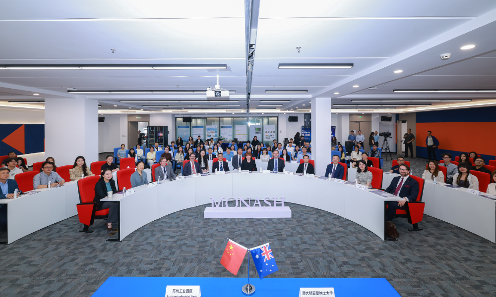 Photograph of a large group of people seated around a curved conference table in a modern meeting room, suggesting a formal gathering or conference. The room features red chairs, white walls with blue and orange accents, and a carpeted floor, with attendees dressed in business attire indicating a professional event.