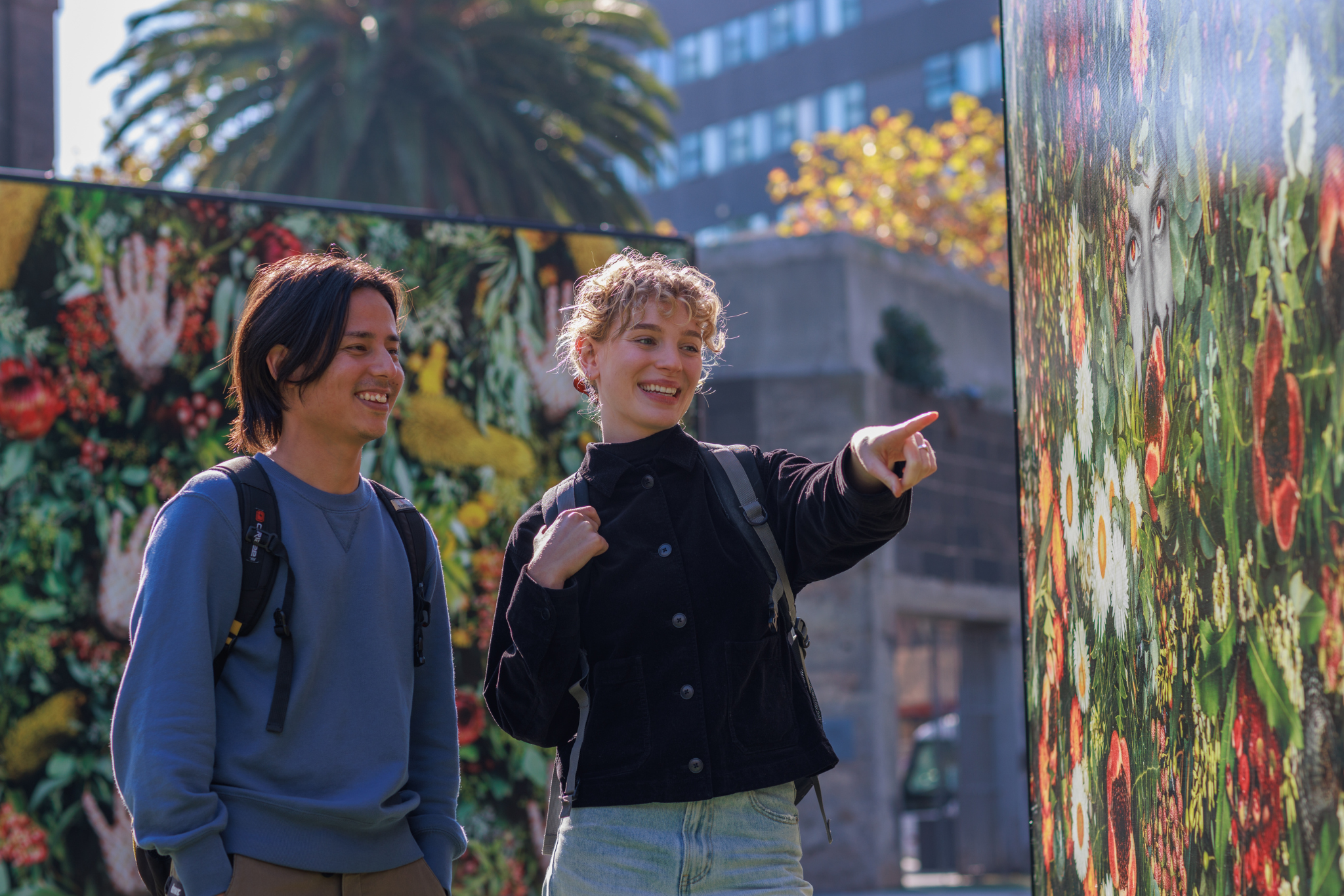 Photograph of two people standing outdoors near large panels covered with colorful floral and nature-themed artwork. One person points towards the artwork while the other looks on, with palm trees and urban buildings visible in the background.