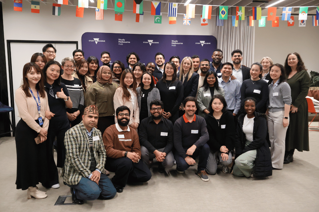 Photograph of a diverse group of people posing together in a conference room with a purple backdrop displaying "Study Melbourne" logos. Group includes men and women of various ages and ethnicities, some seated in front and others standing behind, all smiling and wearing name tags.
