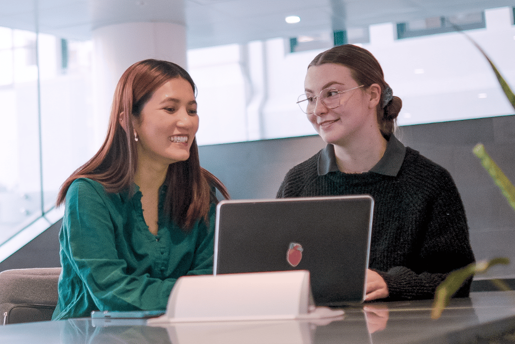 Photograph showing two women engaged in a conversation while looking at a laptop screen. The setting appears to be a modern office or workspace with natural light and a casual atmosphere.