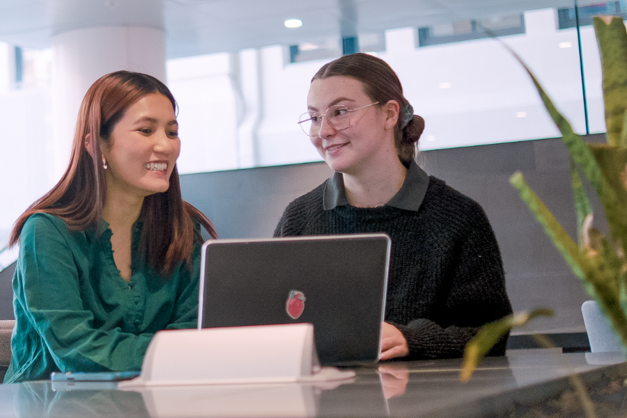 Two people smiling and talking at a desk in a modern office, with laptops open in front of them and a glass wall behind them.