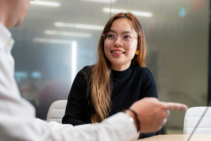 Photograph showing smiling woman with glasses and black turtleneck speaking with partially visible colleague in modern office.