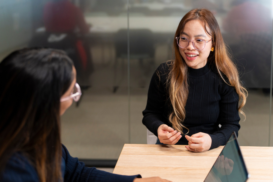Photograph showing smiling woman with glasses and black turtleneck speaking with partially visible person in modern office.