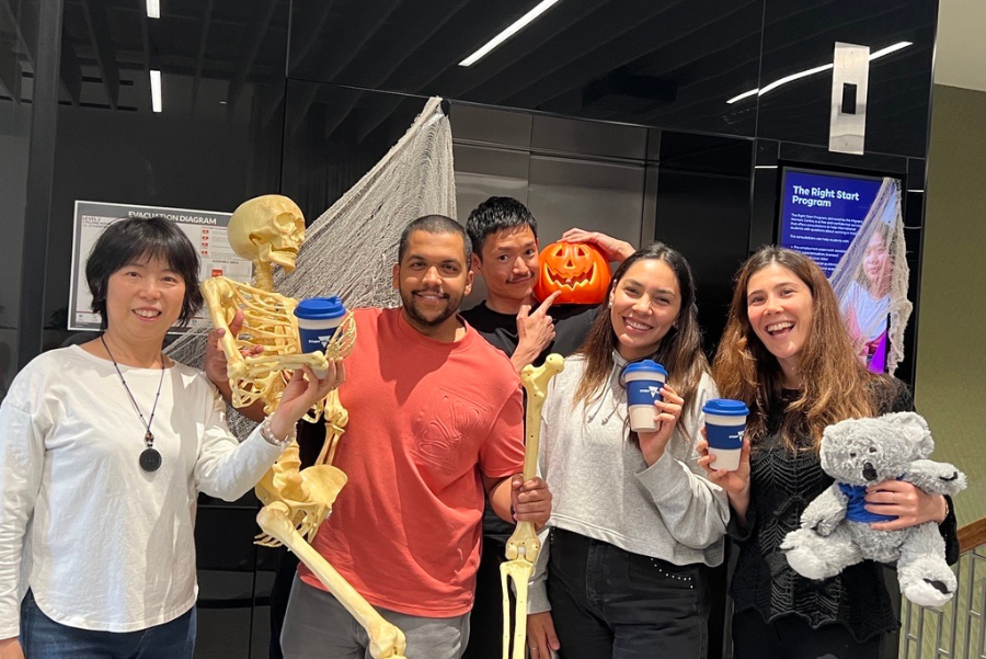 Photograph shows five smiling people posing in a science museum beside a full-size skeleton, with two people holding blue cups, one holding a pumpkin, and another holding a gray stuffed koala. 