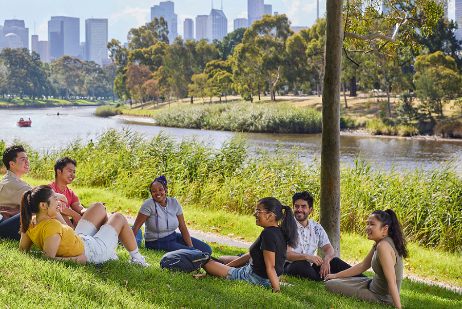 Photograph of a group of seven people sitting on grass near a riverbank with a city skyline in the background. The scene highlights a relaxed outdoor gathering with trees, walking paths, and calm water under a partly cloudy sky.