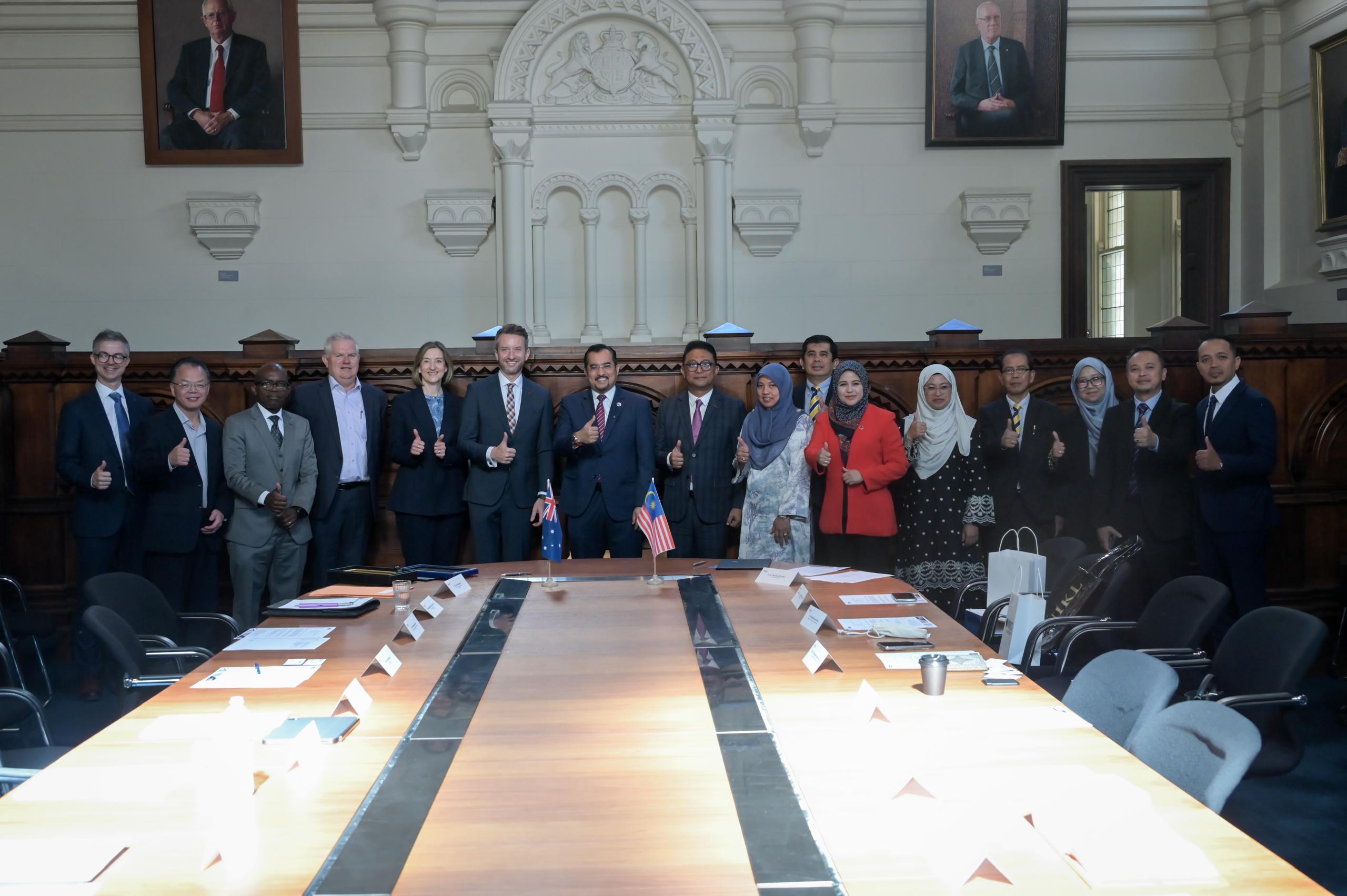 Photograph of a diverse group of professionals standing behind a conference table in a formal meeting room, posing with thumbs up. 