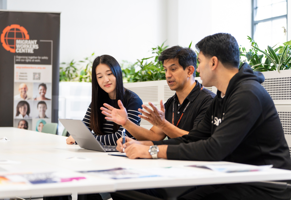 Photograph showing three people engaged in a discussion at a table, with one person holding a tablet and another gesturing with hands