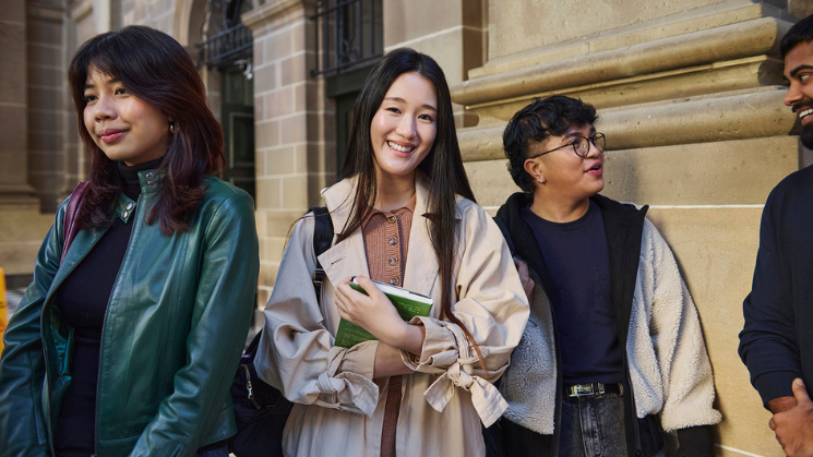 Photograph of four people standing and conversing outdoors against a stone building wall. Three individuals wear casual jackets while one holds a green notebook, suggesting a casual or academic setting.