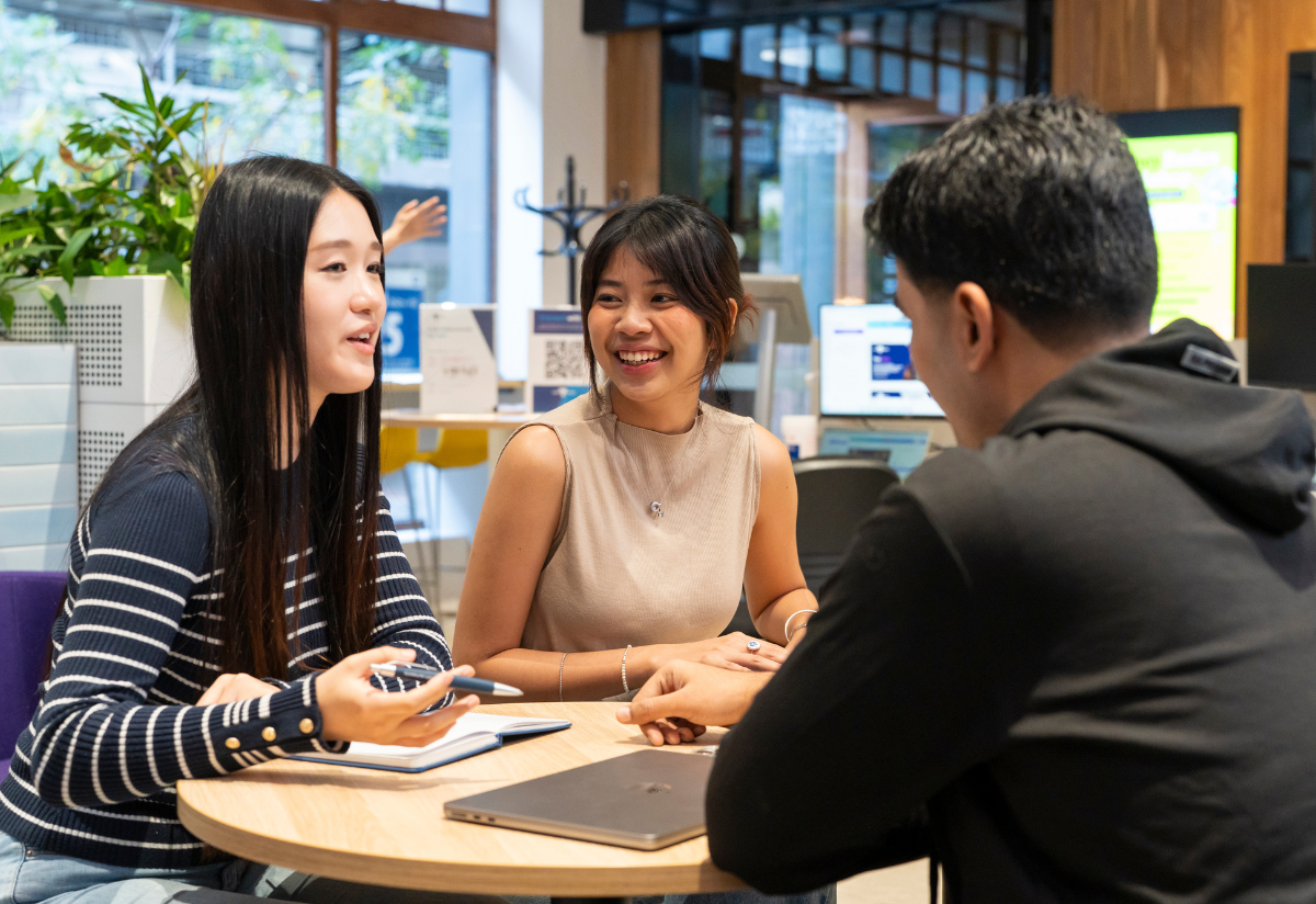 Photograph showing three people engaged in a discussion inside a modern office with computers and plants in the background. The setting suggests a collaborative work environment with natural light and casual attire.