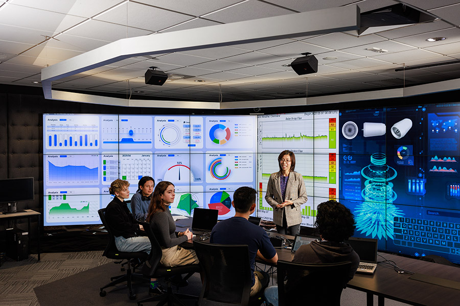 Presenter in meeting, screens with charts and graphs showing behind