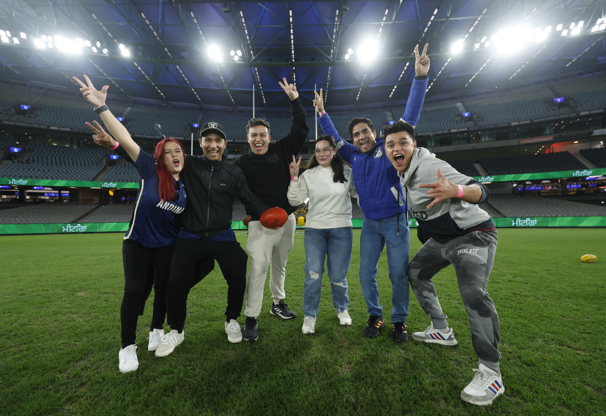 Photograph of six people posing together on a sports field inside a large stadium with bright overhead lights. 