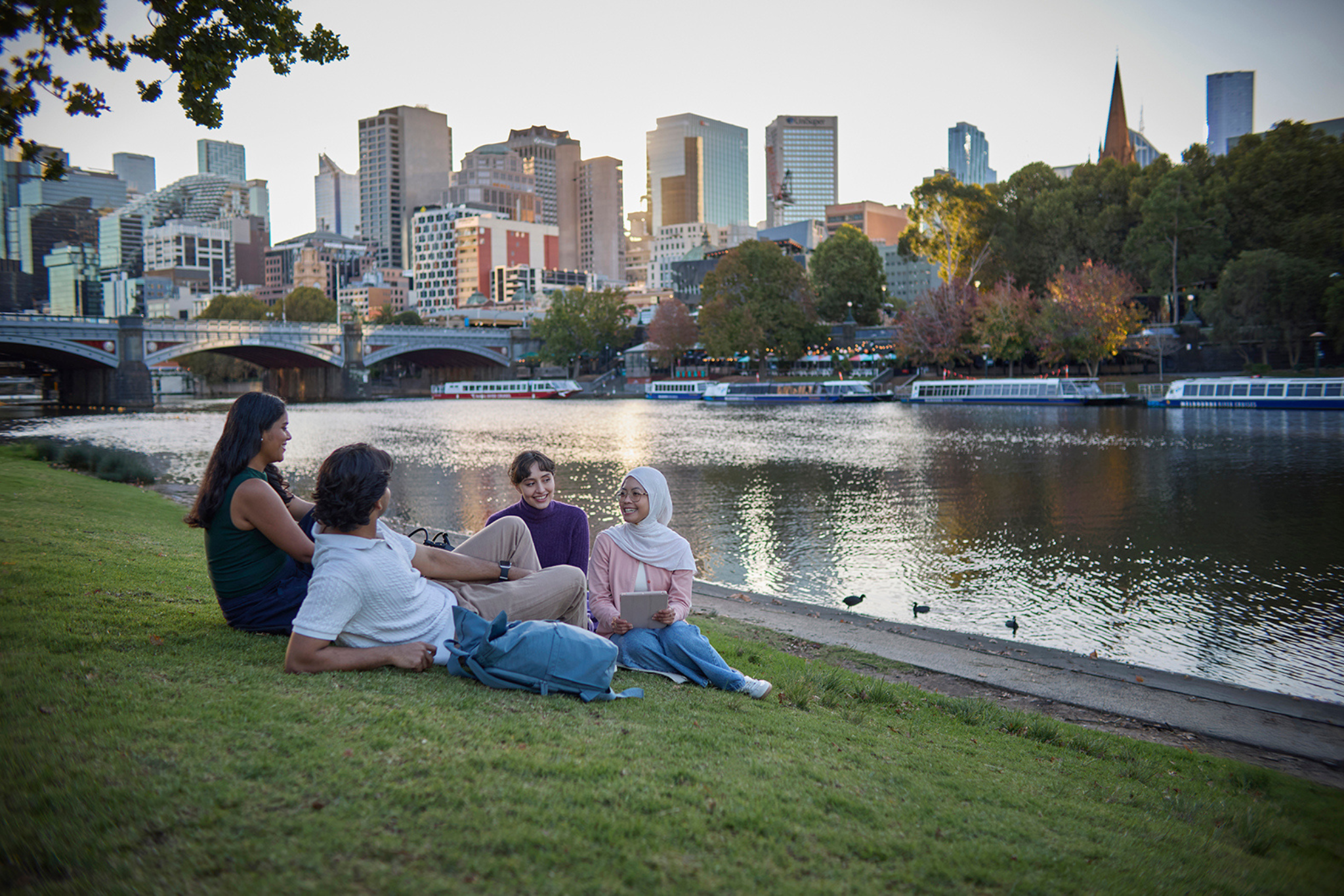 Photograph of four young adults sitting on grass beside a river, engaged in conversation and smiling. Background includes a bridge, boats on water, and autumn-colored trees, suggesting a relaxed outdoor social setting.