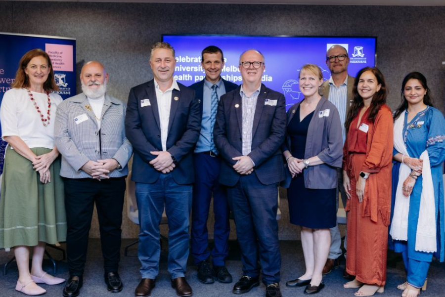 Group of people standing together at a conference stage in front of a blue screen, wearing name badges and semi-formal attire.