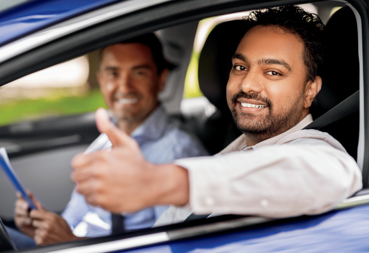 Photograph of two men sitting inside a blue car, with the man in the driver's seat giving a thumbs-up gesture. 