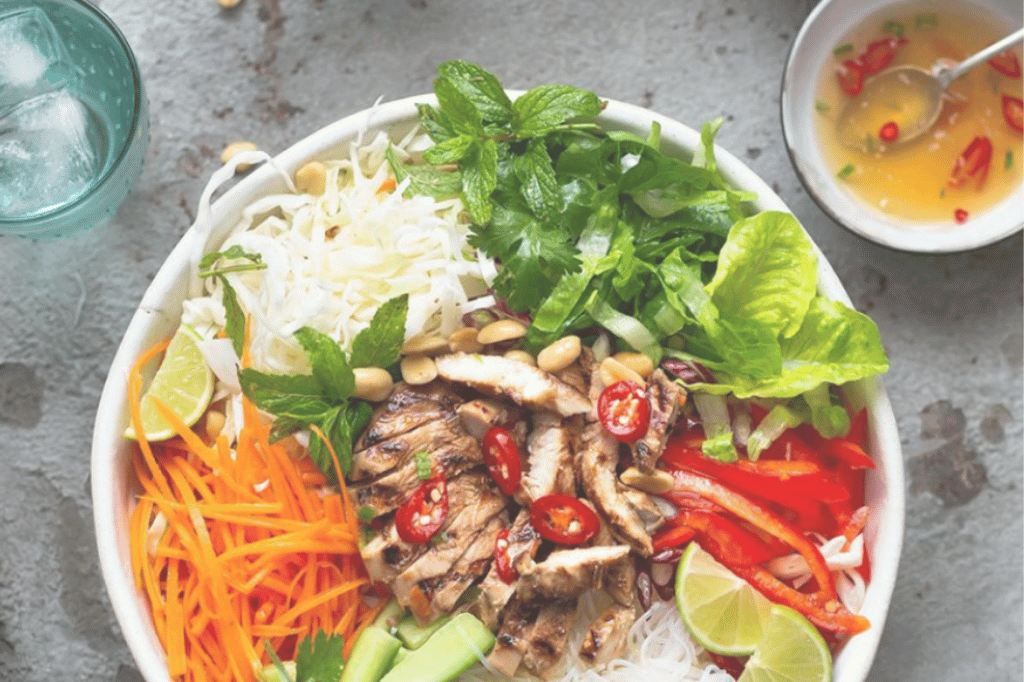 Photograph of a colorful Vietnamese-style rice noodle bowl featuring grilled meat, sliced red chili peppers, shredded carrots, cucumber, lettuce, fresh herbs, peanuts, and lime wedges. The dish is arranged in a white bowl on a gray surface, showcasing a vibrant and fresh meal presentation.