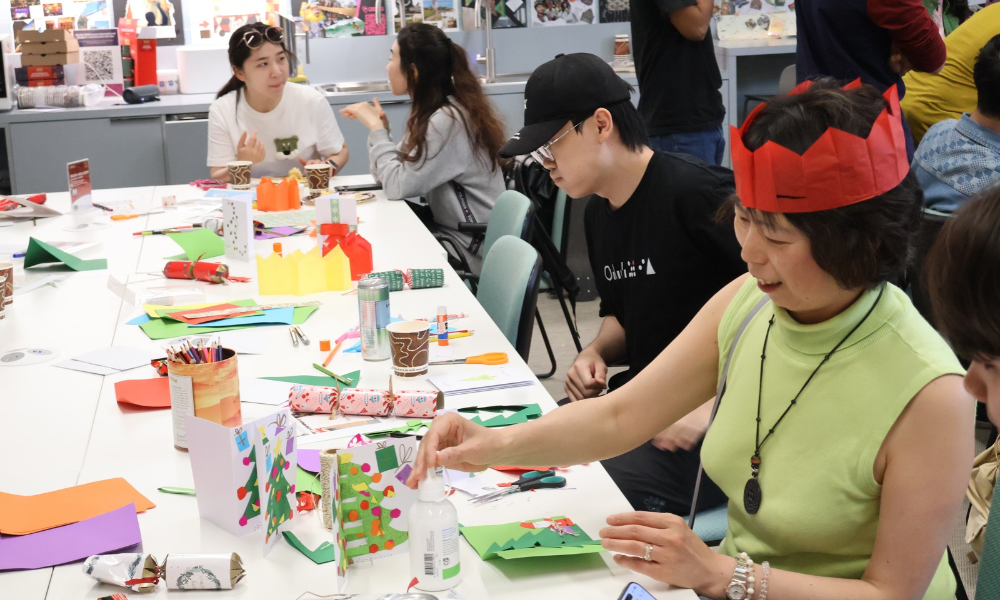 Photograph shows a hands-on arts-and-crafts workshop with participants creating colorful paper projects around a long table. 