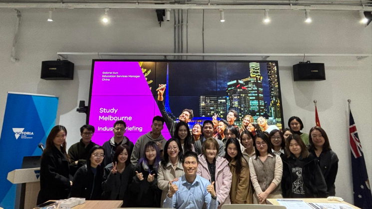 Photograph of a group of people posing indoors at a Study Melbourne event, with a large purple presentation screen and a Victoria state banner visible in the background. 