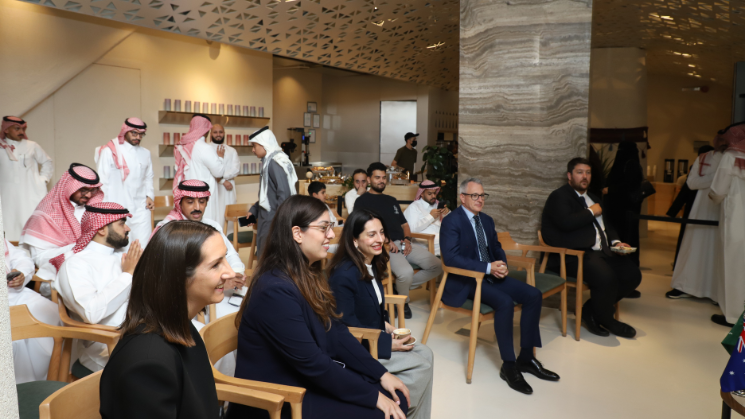 Photograph of a formal gathering or meeting with attendees seated and listening attentively. The setting includes men and women dressed in business and traditional attire, arranged in rows with a speaker or presenter standing on the right side.