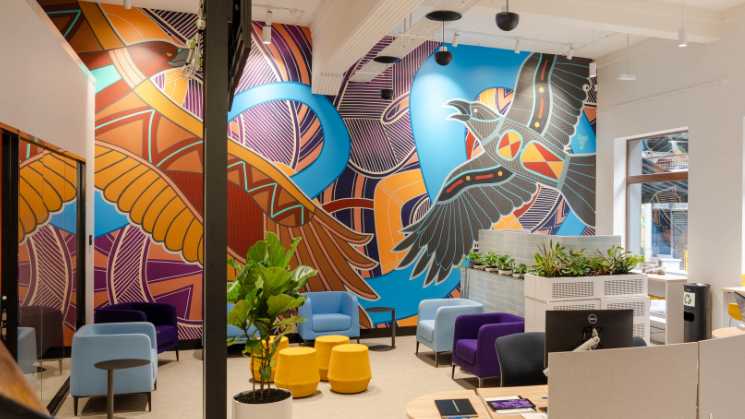 Photograph of a modern office lounge featuring colorful seating including blue, purple, and yellow chairs and ottomans arranged around a central plant. Walls display a large, vibrant mural with abstract patterns and a stylised bird in shades of orange, blue, black, and red. 
