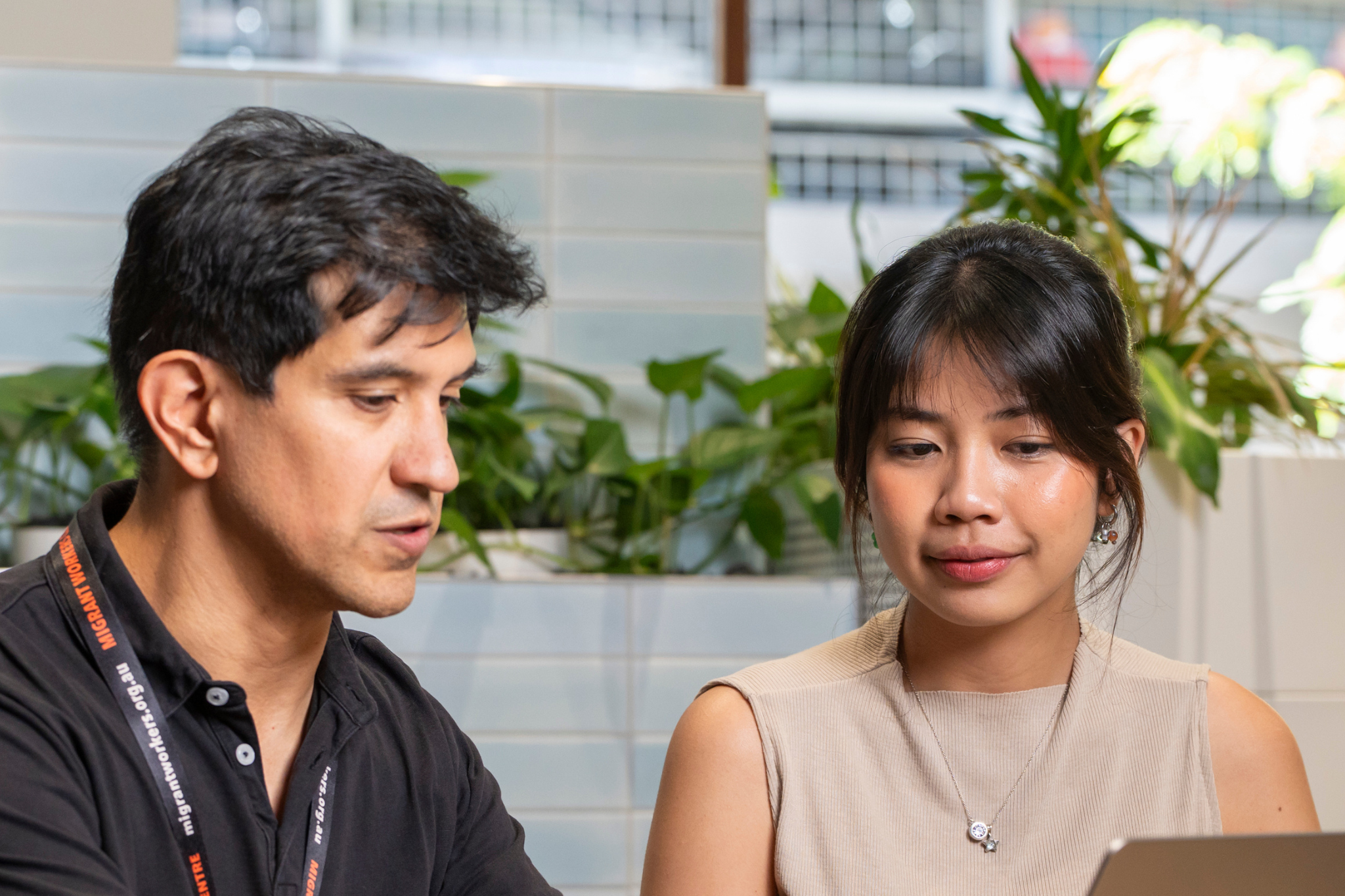 Photograph of two people engaged in a discussion in an indoor setting with plants and tiled walls in the background. The man wears a black shirt with a lanyard, and the woman wears a sleeveless beige top, both focused on a task.