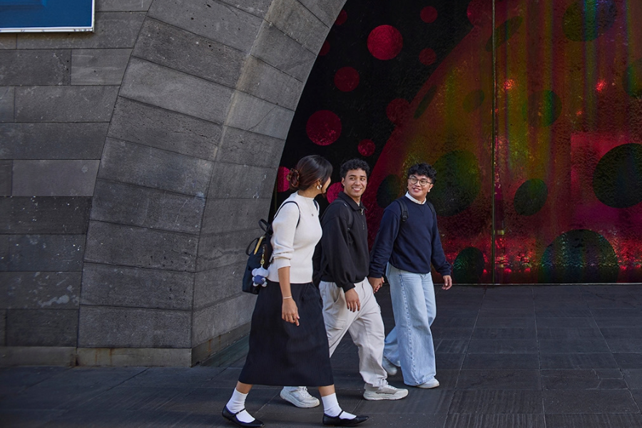 Photograph shows three people walking past a stone archway and vibrant abstract public art installation. 