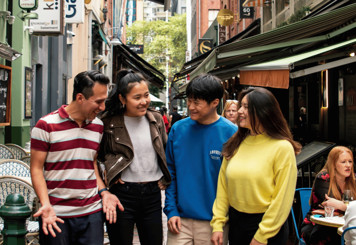 Photograph of four people standing and talking outdoors on a narrow street with shops and greenery in the background. 