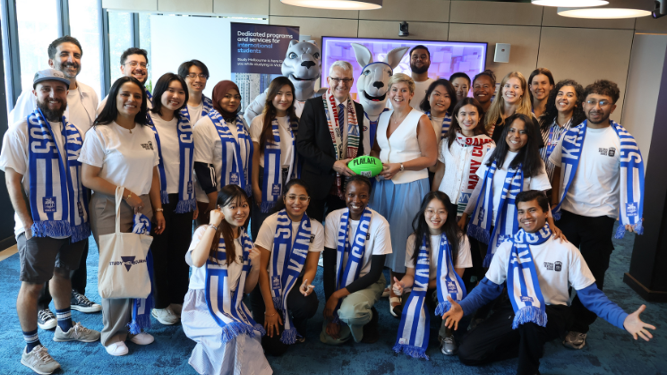 Photograph of a diverse group of people wearing blue and white scarves, posing indoors with two individuals dressed in mascot costumes.