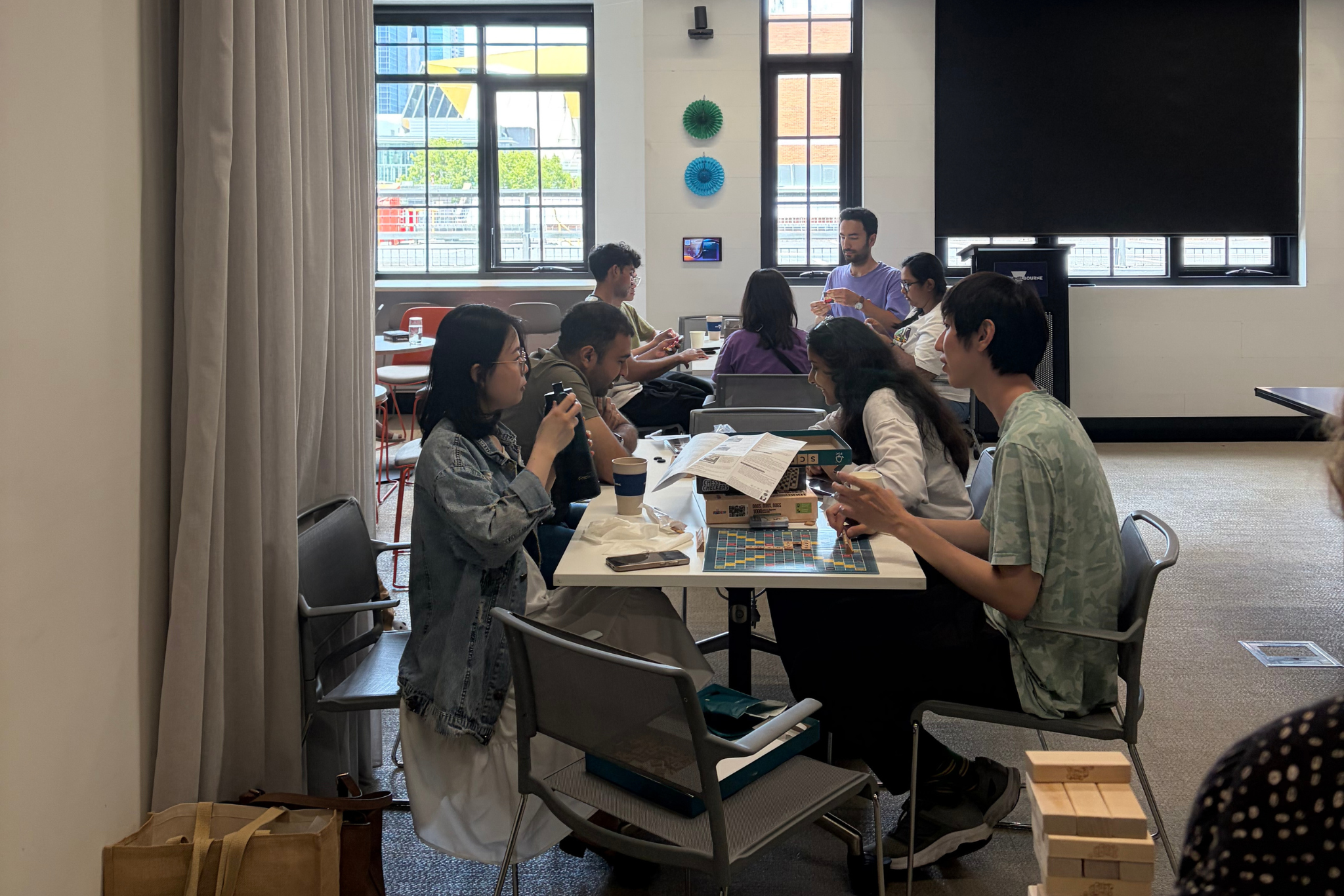 Photograph of a group of people seated around a table in a casual indoor setting, engaged in conversation and activities like drinking coffee and using phones. The room features large windows with natural light, modern decor with colorful wall hangings, and a relaxed atmosphere suggesting a social or collaborative gathering.