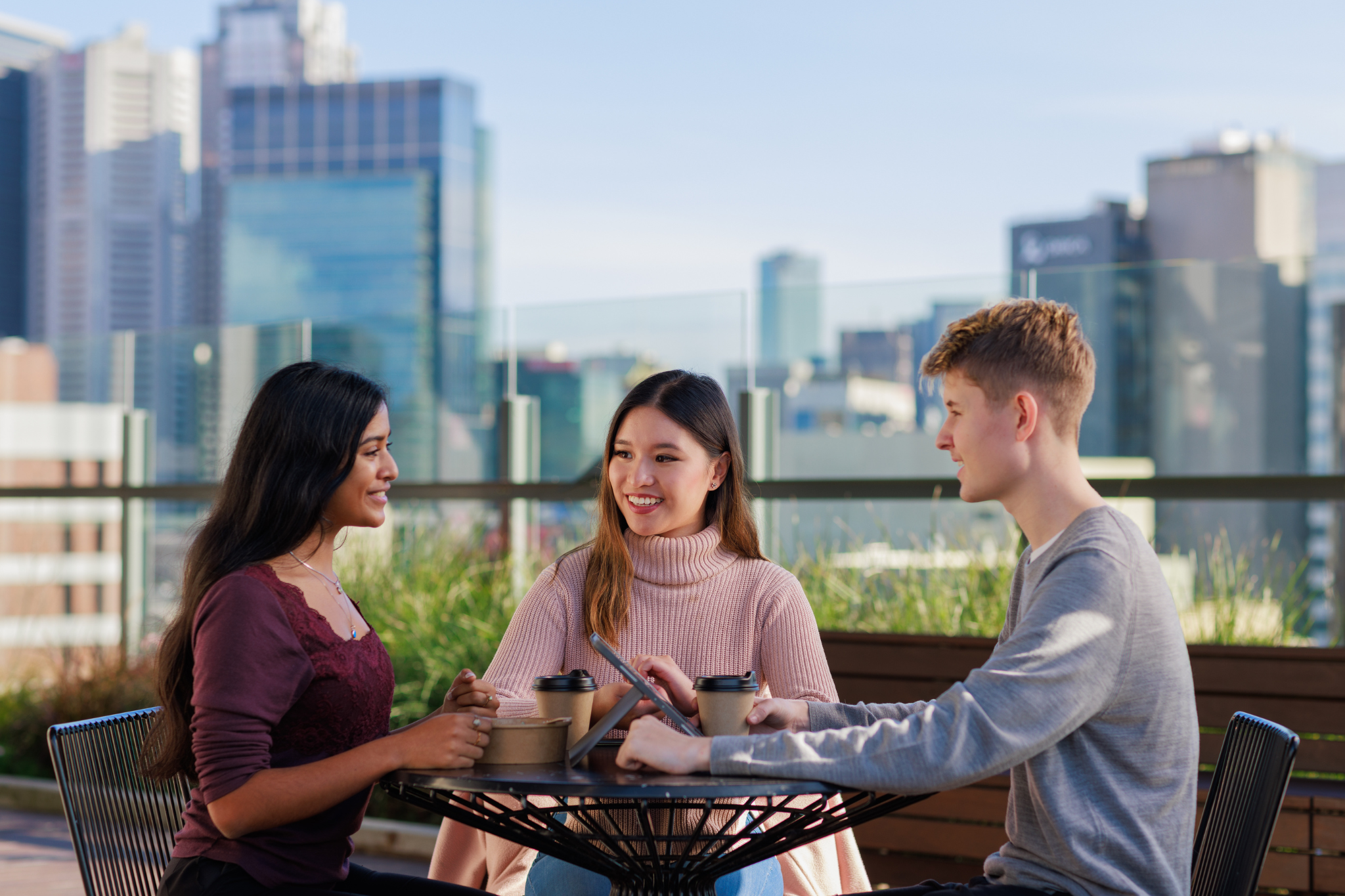 Photograph of three people sitting outdoors at a table with coffee cups, engaged in conversation. Background shows urban buildings and greenery, suggesting a casual meeting or social gathering in a city setting.