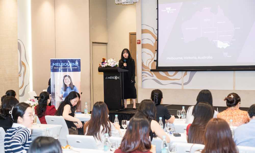 Photograph of a conference presentation showing a speaker at a podium addressing an audience seated at tables. 
