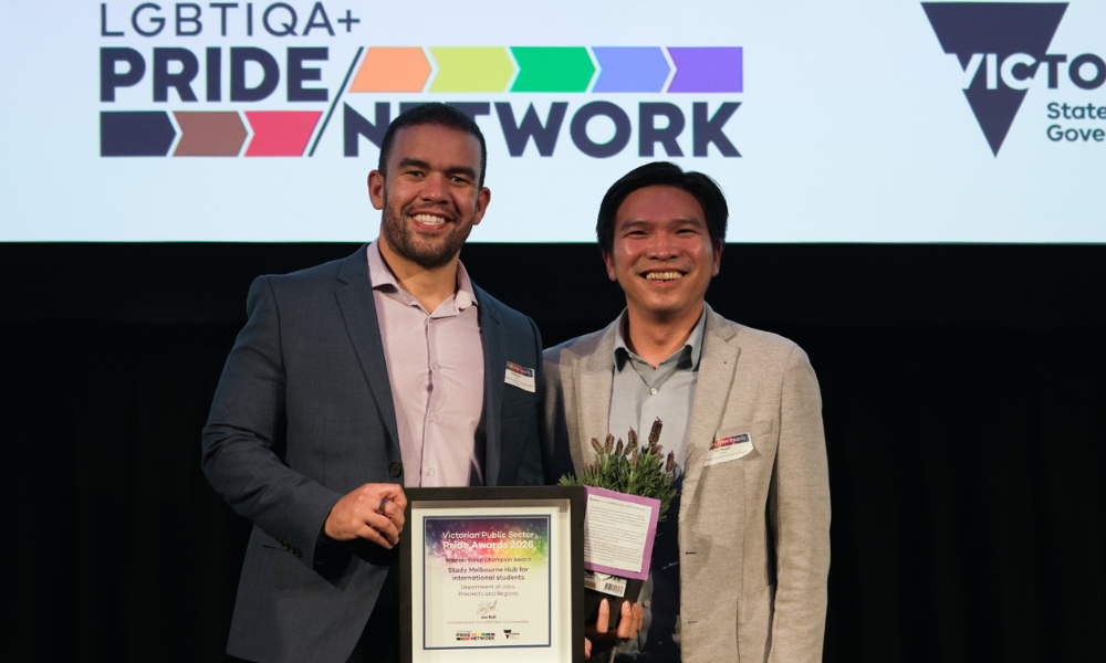 Photograph of two men standing side by side on a stage, holding a framed certificate and a small bouquet of flowers. 