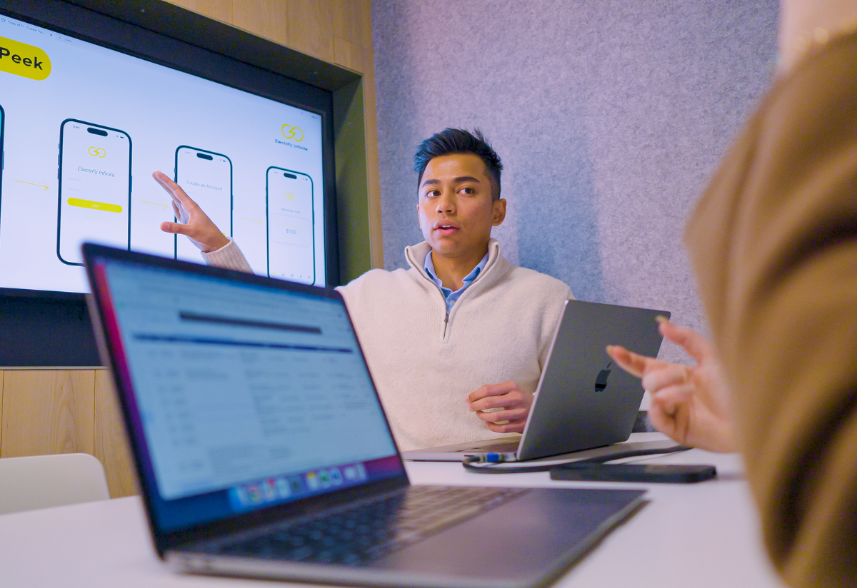 Photograph of a person presenting app interface designs displayed on a large screen, showing different smartphone layouts with color-coded elements in blue, yellow, and white. Two other participants are visible with laptops, one pointing towards the presenter, indicating a collaborative discussion. 