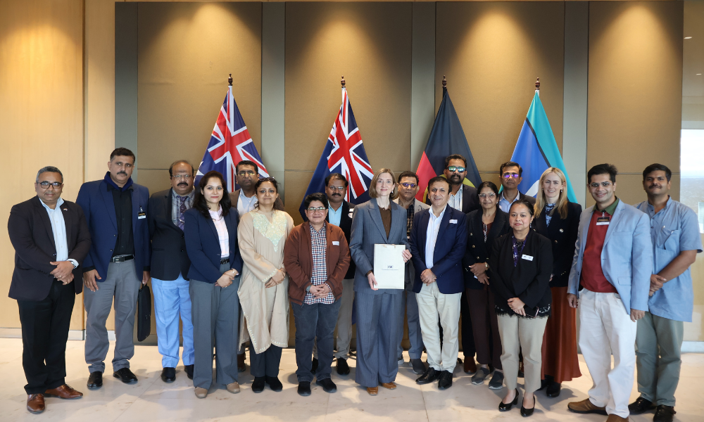 Photograph of a diverse group of people standing indoors in front of four flags, including two Union Jack flags, a black and red flag, and a blue and green flag. The group appears to be participating in a formal or official event, with one person holding a certificate or document at the center.