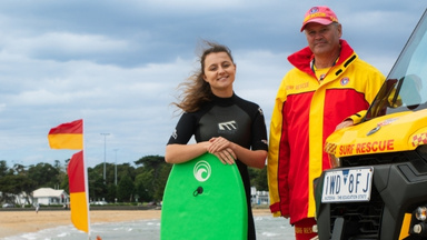 International student with boogie board standing next to surf lifesaver, red and yellow flags and beach in the background