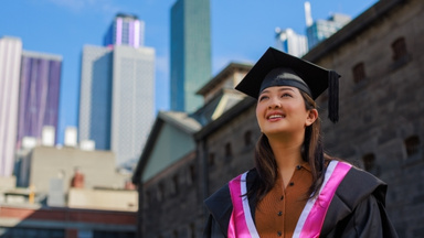 Graduate in gown and mortar board, city buildings in background