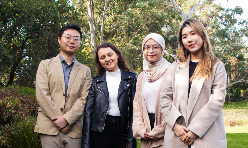 Photograph of four diverse adults posing together in a sunny, leafy park. 