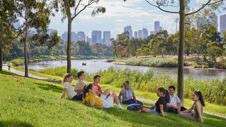 Students enjoying summer on the banks of the Yarra River, city skyline in background