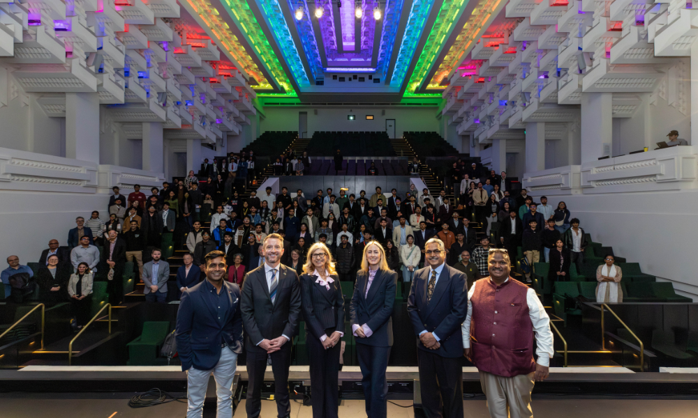 Photograph of a large group of people gathered in an auditorium with five individuals standing in front, dressed in formal and business attire. The setting suggests a professional event or conference, with attendees seated in tiered rows and a well-lit stage area.