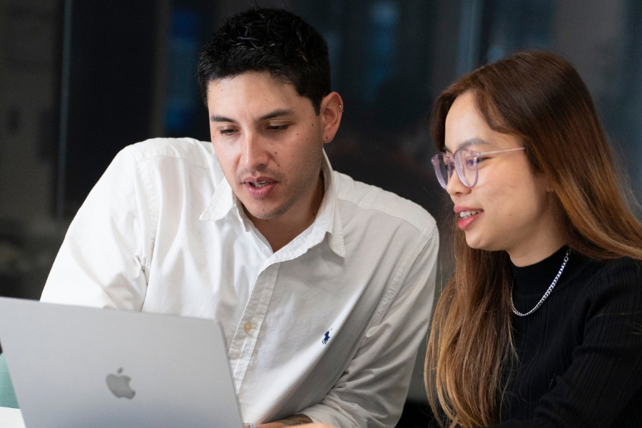 Photograph showing two coworkers collaborating while viewing a silver Apple laptop. 