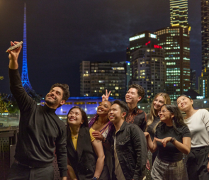 A group of students taking a selfie together at night time, with the Melbourne skyline in the background.
